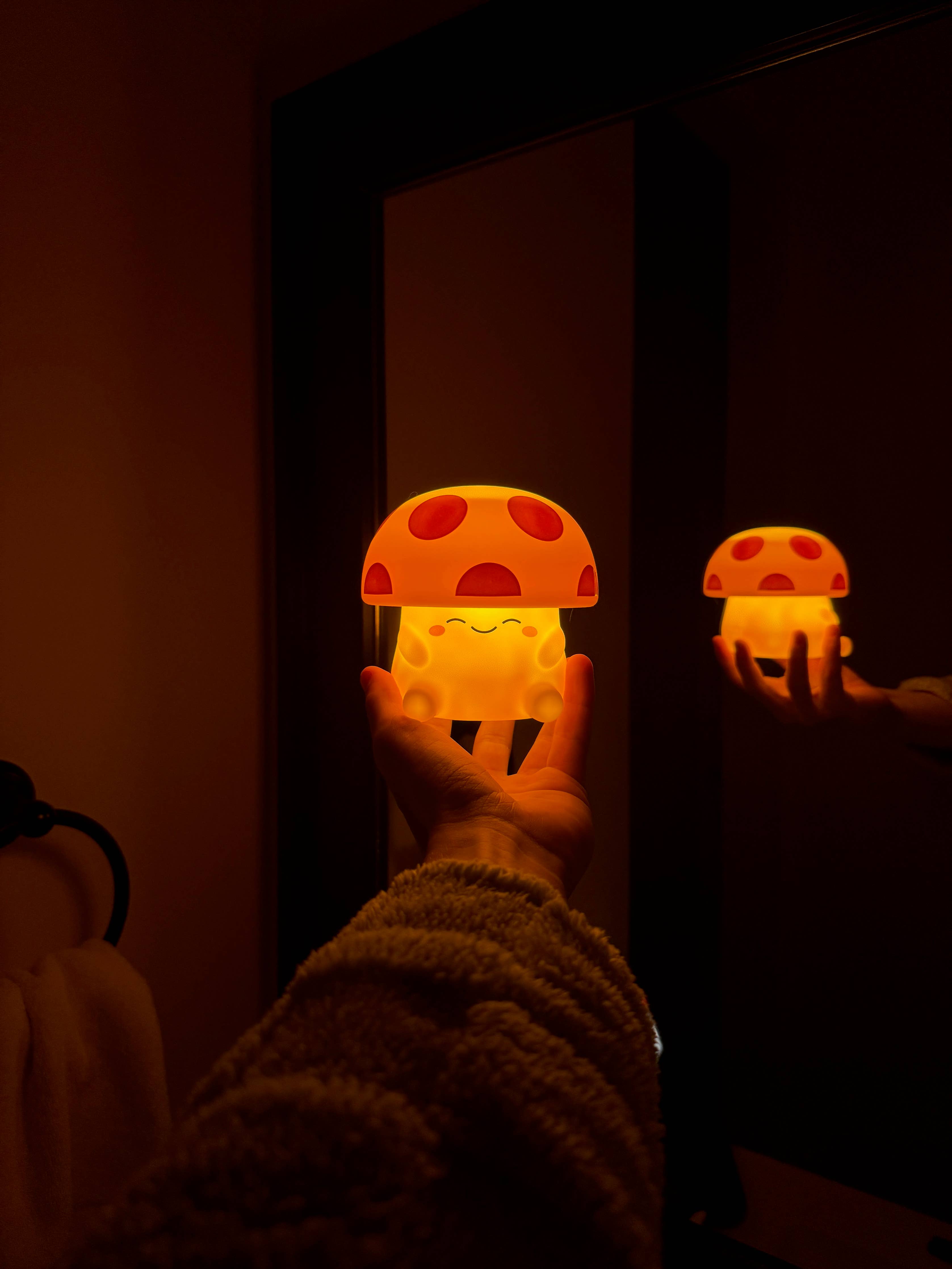 Hand holding a glowing Mushi mushroom night light in front of a mirror in a cozy warm-lit room